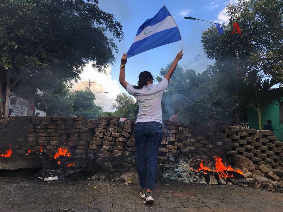 Mujer de espaldas levantando la bandera de Nicaragua frente a una barricada de adoquines con llantas encendidas