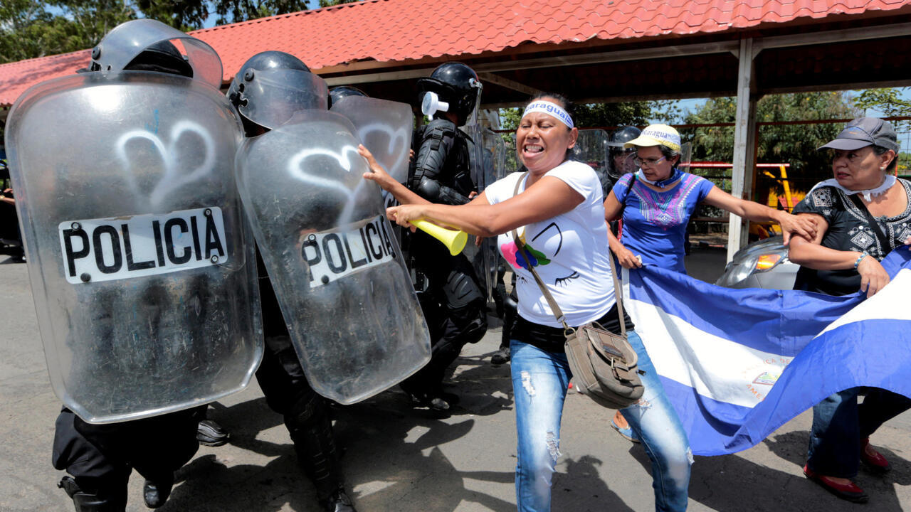 Mujeres con la bandera de Nicaragua reclamando frente a policías con escudos antimotines