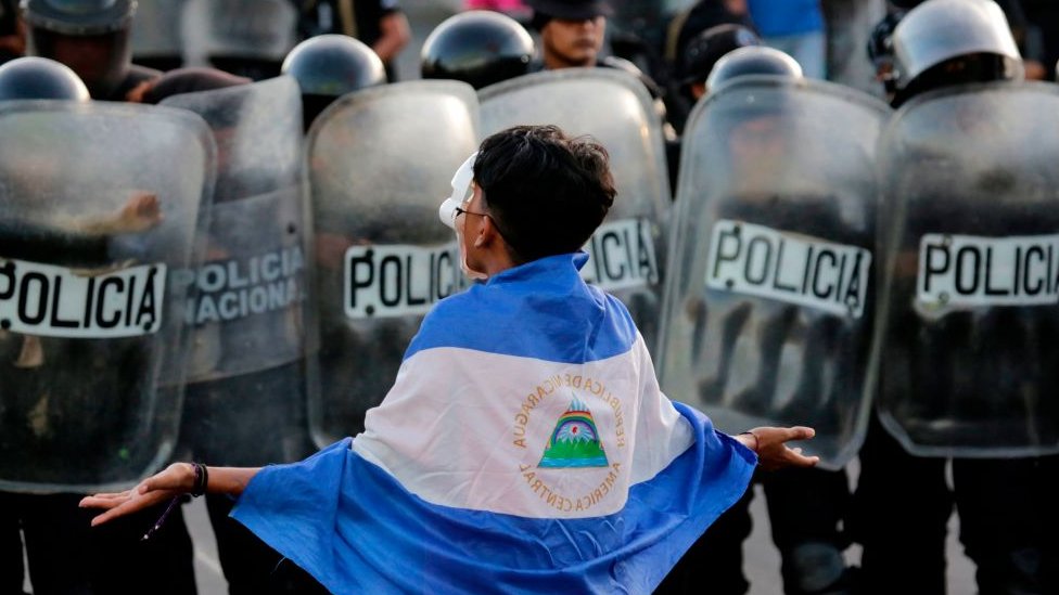 Persona envuelta en la bandera de Nicaragua frente a una fila de escudos de la Policía