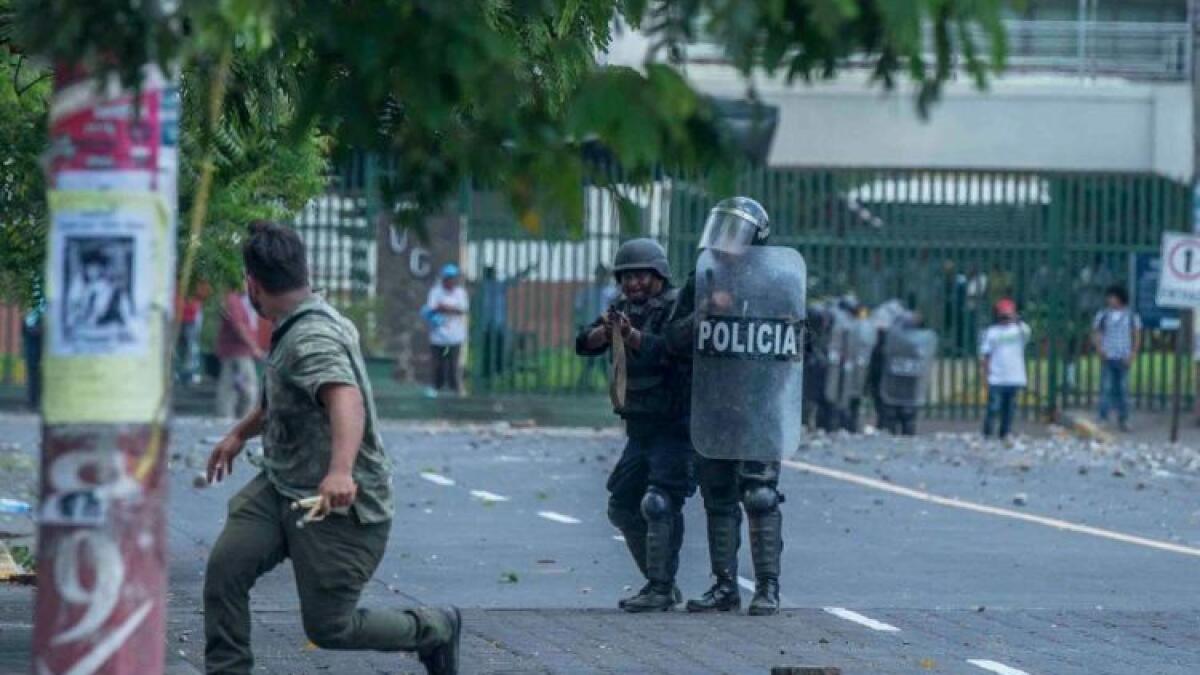 Manifestante lanzando una piedra ante policías antimotines que apuntan con un arma